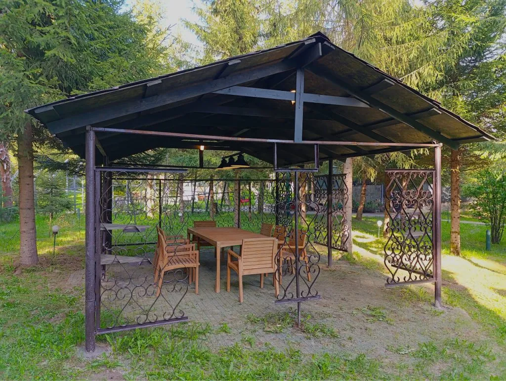 Уличная беседка с деревянным столом и креслами в зеленом парке, окруженная деревьями и газоном. Идеальное место для отдыха на природе в ВЕ Art Resort в Подмосковье.